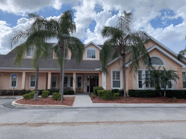 Front exterior view of a single-story building with peach-colored walls, white trim, and a gray shingled roof. The entrance is centered with a covered porch supported by columns. Several palm trees and shrubs are planted in mulched beds along the front. The sky is partly cloudy.