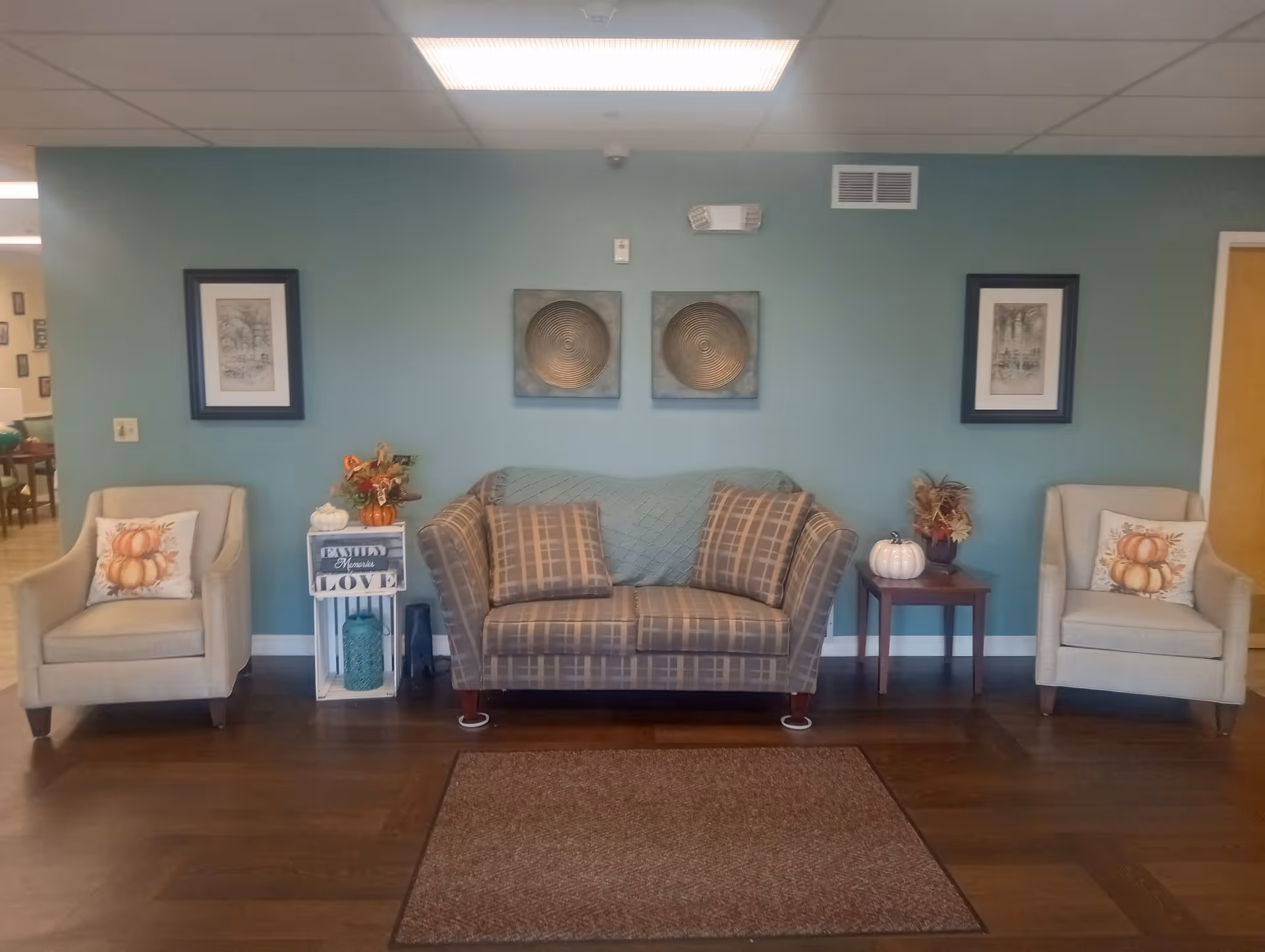 A cozy sitting area with a plaid loveseat in the center flanked by two beige armchairs with pumpkin-themed pillows. The wall behind is painted teal and decorated with two framed pictures and two circular metallic wall art pieces. There are small tables on either side of the loveseat with decorative items including white pumpkins and floral arrangements. The floor is wooden with a brown rug in front of the seating.