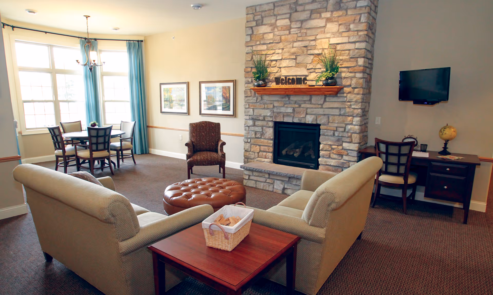 A cozy living room area with two beige sofas facing each other, a round brown leather ottoman in the center, and a stone fireplace with a wooden mantle decorated with plants and a welcome sign. To the left, there is a dining table with six chairs near large windows with blue curtains. On the right side, a small desk with a chair, a globe, and a wall-mounted TV are visible. The room has beige walls and carpeted flooring.
