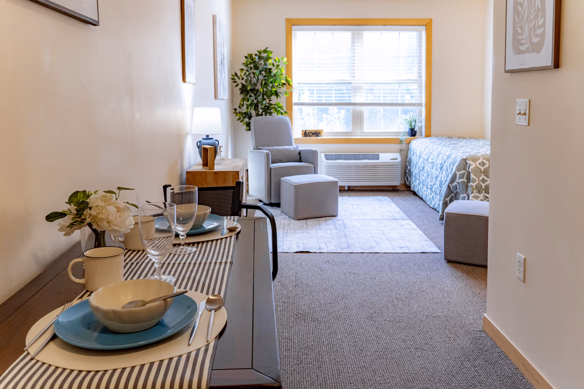 A cozy studio apartment with a dining table set for two on the left, featuring plates, bowls, glasses, and a small flower arrangement. In the background, there is a comfortable armchair with an ottoman, a small side table with a lamp, a window with blinds, and a single bed with a patterned bedspread. The room has neutral-colored walls and carpeted flooring.