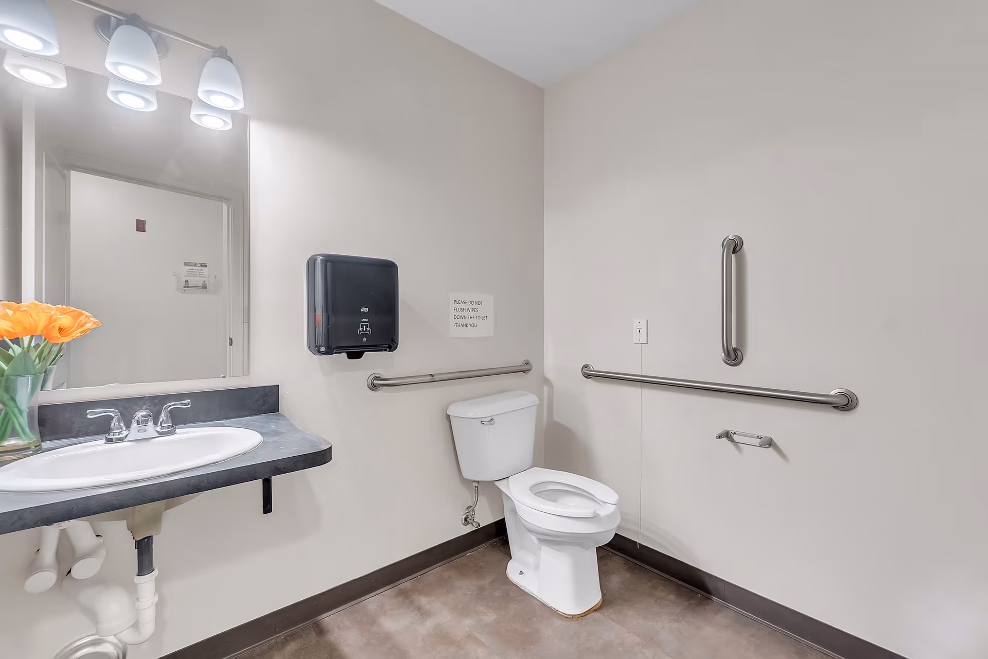 Accessible restroom with a toilet and grab bars, a sink and mirror, paper towel dispenser, and a vase of orange flowers on the counter.