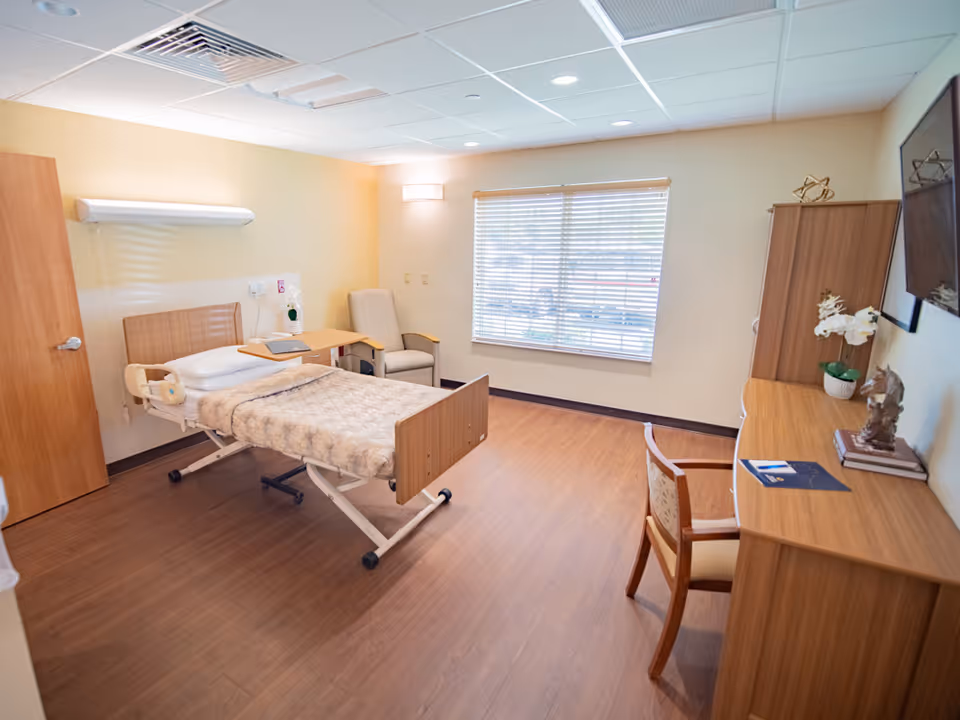 A clean and well-lit bedroom in a healthcare facility featuring a hospital bed with a beige blanket, a beige armchair, a wooden desk with a chair, decorative items including a plant and books, a wall-mounted TV, and a window with blinds letting in natural light.