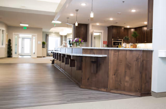 Interior view of a senior living facility showing a long kitchen island with a granite countertop and several wooden stools. The kitchen area has dark wood cabinets, built-in ovens, and pendant lights hanging from the ceiling. In the background, there is a hallway leading to a glass door entrance with natural light coming through.