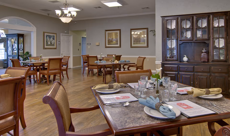 A dining room in Churchill Terrace Senior Living with several wooden tables and chairs arranged neatly. Tables are set with plates, glasses, napkins, and menus. The room has wooden flooring, framed artwork on the walls, a chandelier, and a large wooden cabinet displaying plates.