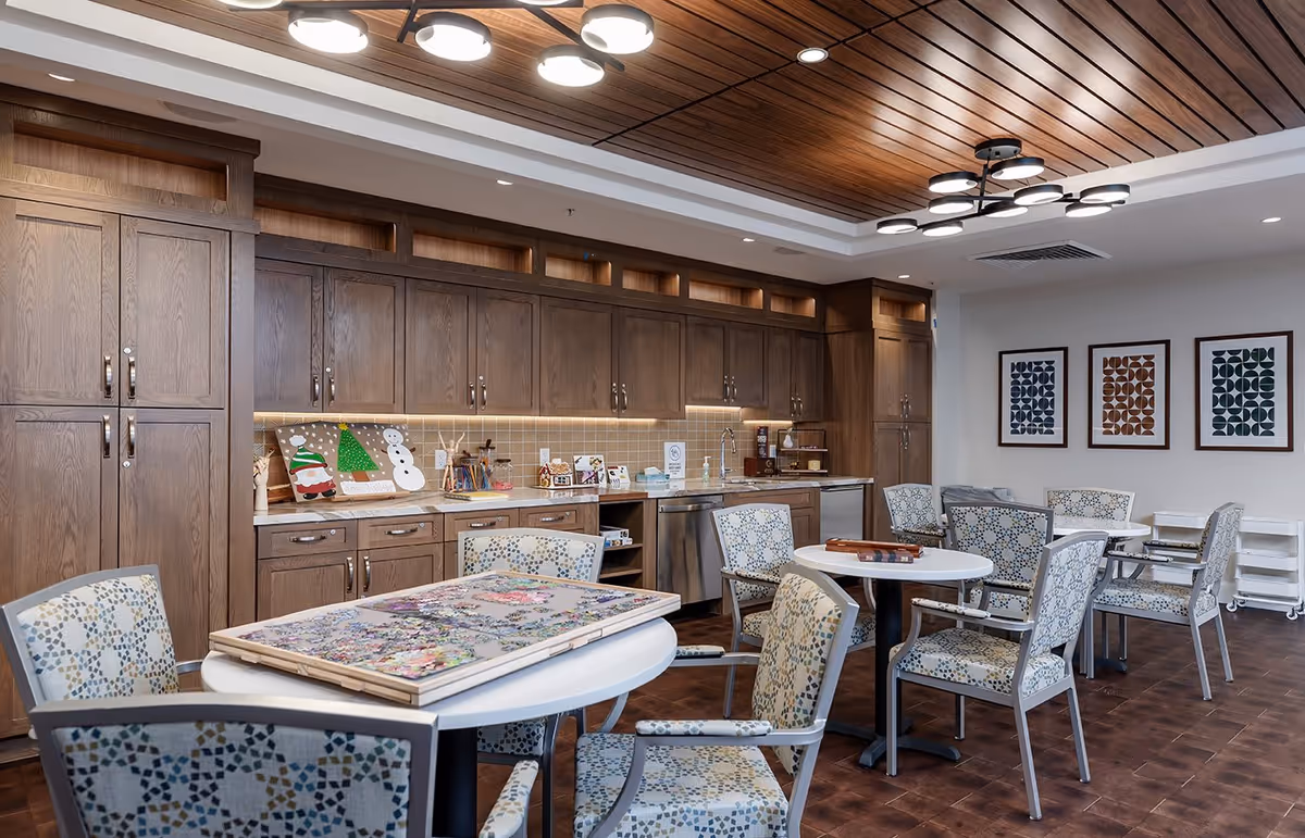 A spacious activity room with wooden cabinets and a countertop along one wall. Several round tables with patterned chairs are arranged in the room, one table has a large puzzle in progress. The ceiling features wooden paneling and modern circular light fixtures. Three framed abstract artworks hang on the white wall.