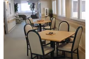 A bright interior room with several square wooden tables and cushioned chairs arranged around them. Large windows line one side of the room, letting in natural light. In the background, there is a seating area with armchairs and a potted plant near the window.