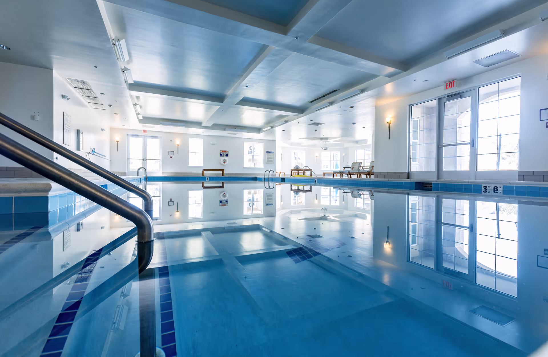 Indoor swimming pool area with clear blue water, stainless steel handrails, and large windows letting in natural light. The ceiling has a grid pattern with recessed lighting, and there are chairs and tables along the far wall near the windows.