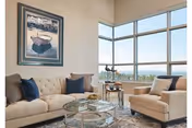 Bright living room with a beige sofa and armchair, glass coffee table, framed artwork, and large corner windows with an outside view.