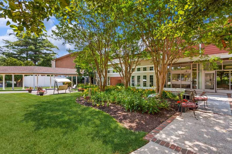 Outdoor garden area at HeartLands Senior Living Village at Ellicott City featuring green grass, trees, flower beds, benches, a covered walkway, and a building with large windows in the background under a blue sky with some clouds.