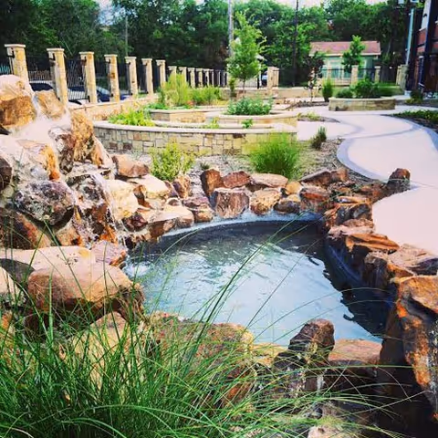 Outdoor garden area with a small pond surrounded by rocks and greenery, a stone retaining wall, a paved walkway, and a fence with stone pillars in the background.