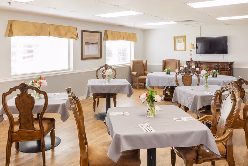 A bright dining room with several tables covered in gray tablecloths, each set with silverware and a small vase of flowers. The room has wooden chairs with ornate backs, two armchairs near the back wall, two windows with beige valances, framed artwork on the walls, and a flat-screen TV mounted above a wooden cabinet.