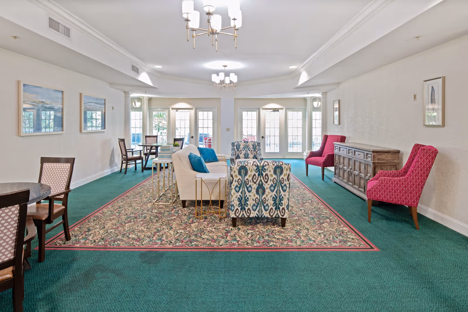 Bright communal lounge with sofas, patterned armchairs, and tables arranged on a large area rug facing French doors.