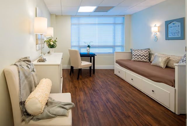 A cozy room with a white daybed featuring multiple decorative pillows, a small black table with a beige chair in front of a large window with blinds, a white dresser with a lamp and flowers, and a cushioned bench with a throw blanket and pillow. The room has wooden flooring and soft lighting from wall sconces and a ceiling light.