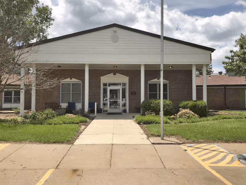 Front exterior view of a single-story brick building with a white covered entrance supported by four white columns. There is a concrete walkway leading to glass double doors, flanked by windows and greenery on both sides. A flagpole is positioned near the walkway, and the sky is partly cloudy.