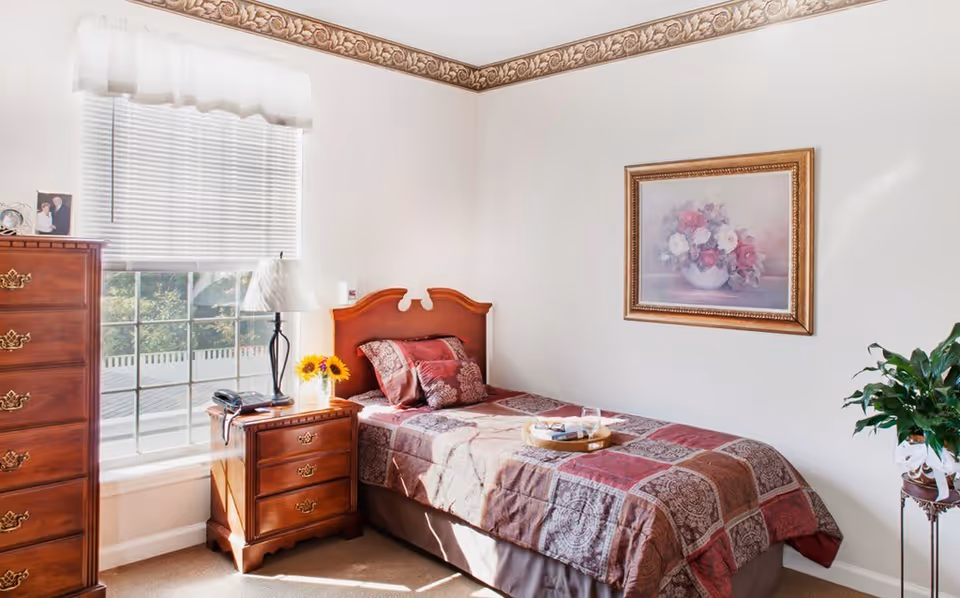 A cozy bedroom in an assisted living facility featuring a single bed with a patterned quilt and pillows, a wooden nightstand with a lamp, a telephone, and a vase with sunflowers. There is a wooden dresser next to a window with blinds and a white valance. A framed floral painting hangs on the wall, and a green plant is placed on a stand in the corner.