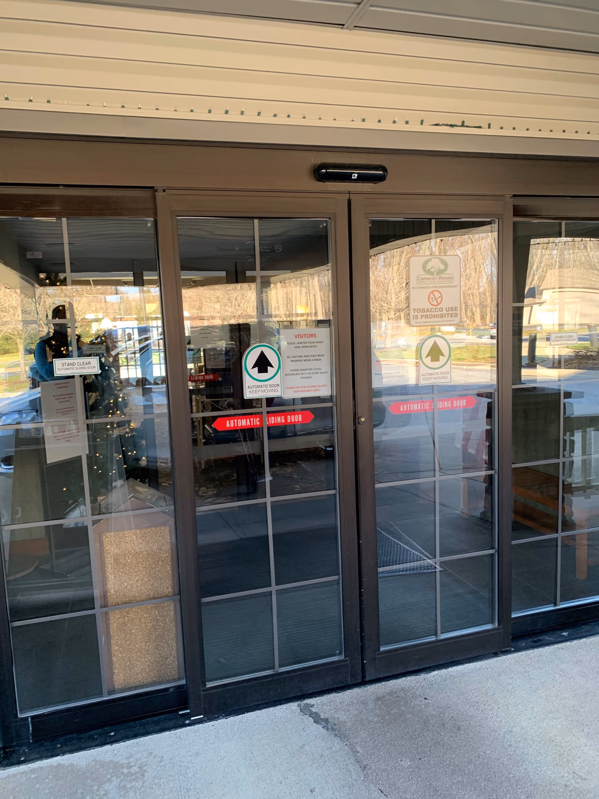Glass automatic sliding entrance doors of a senior living community with signage and outdoor reflections.
