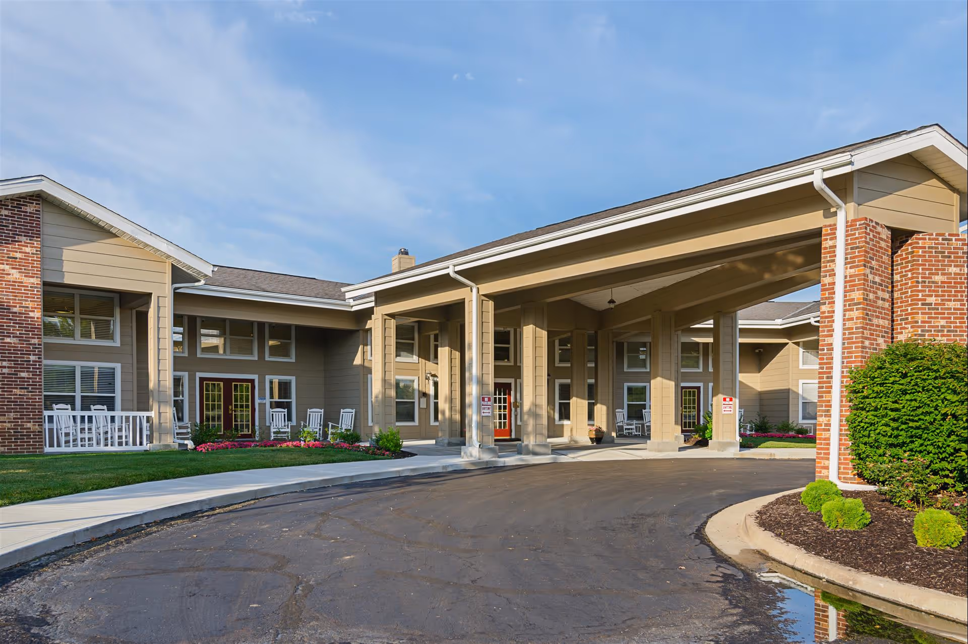 Front entrance of an assisted living facility with a covered porte-cochère, porch rocking chairs, and landscaped beds.