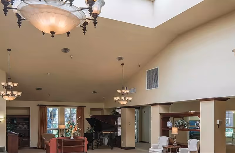 A spacious living room area with high ceilings and multiple hanging light fixtures. The room features a grand piano, a red sofa, two white armchairs, a wooden side table with a lamp, and large windows with curtains allowing natural light to enter.