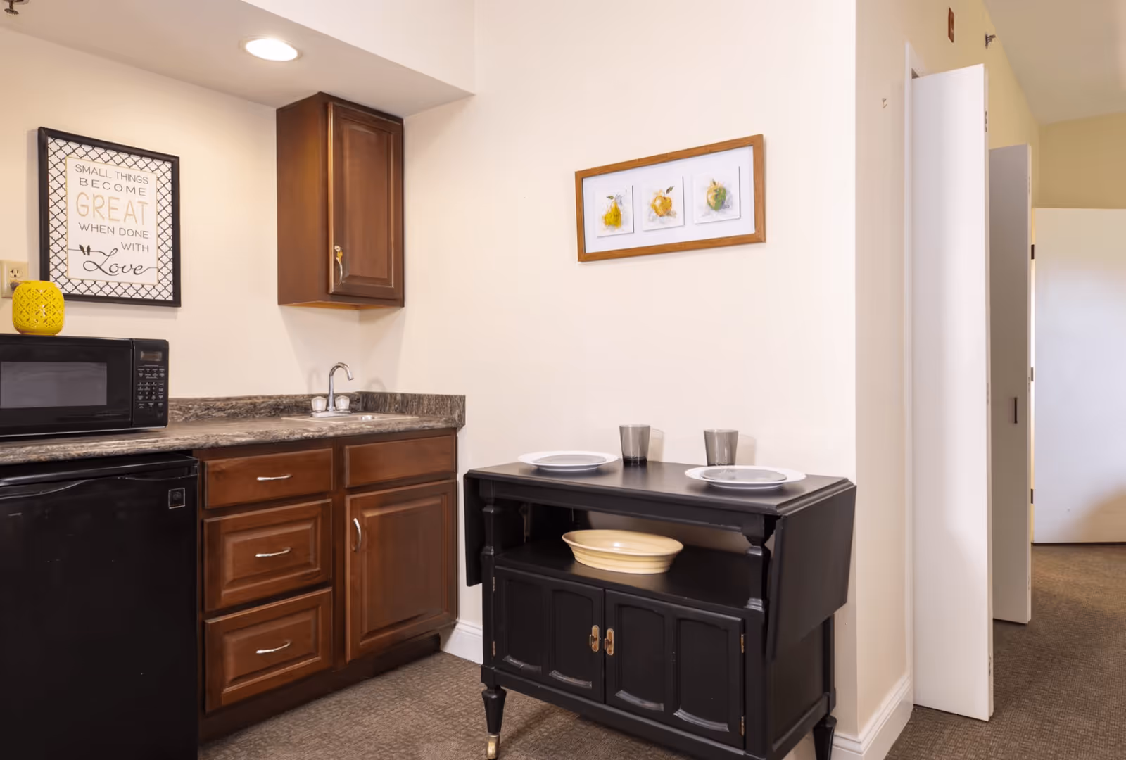 Small kitchenette with dark wood cabinets, a microwave and sink, and a black buffet table set with plates against a beige wall.