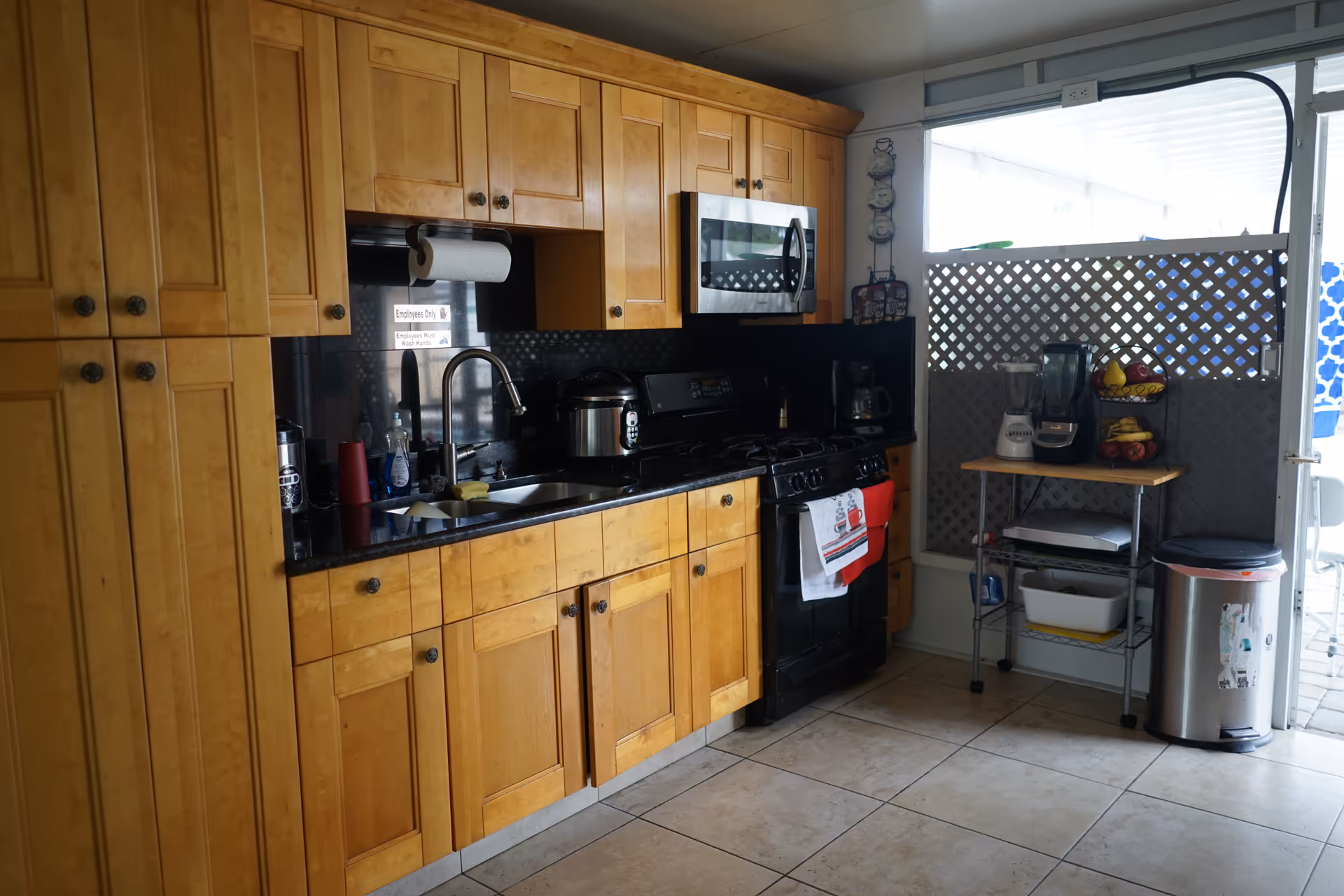 A kitchen with wooden cabinets, a black countertop, a sink with a faucet, a microwave oven mounted above a black stove, and various kitchen appliances including a blender and a coffee maker on a small metal rack. There is a trash can to the right and a window with a lattice screen behind the rack.