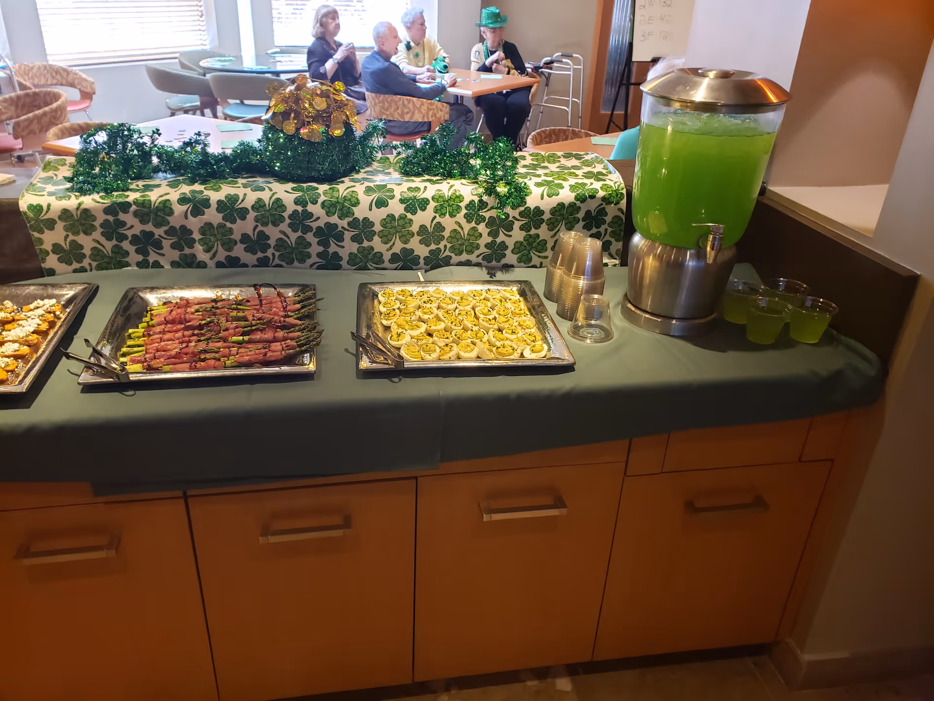 A buffet table with trays of appetizers including bacon-wrapped asparagus and deviled eggs, along with a dispenser of green beverage and plastic cups. The table is decorated with a shamrock-themed cloth and green decorations. In the background, elderly people are seated at tables in a common area.