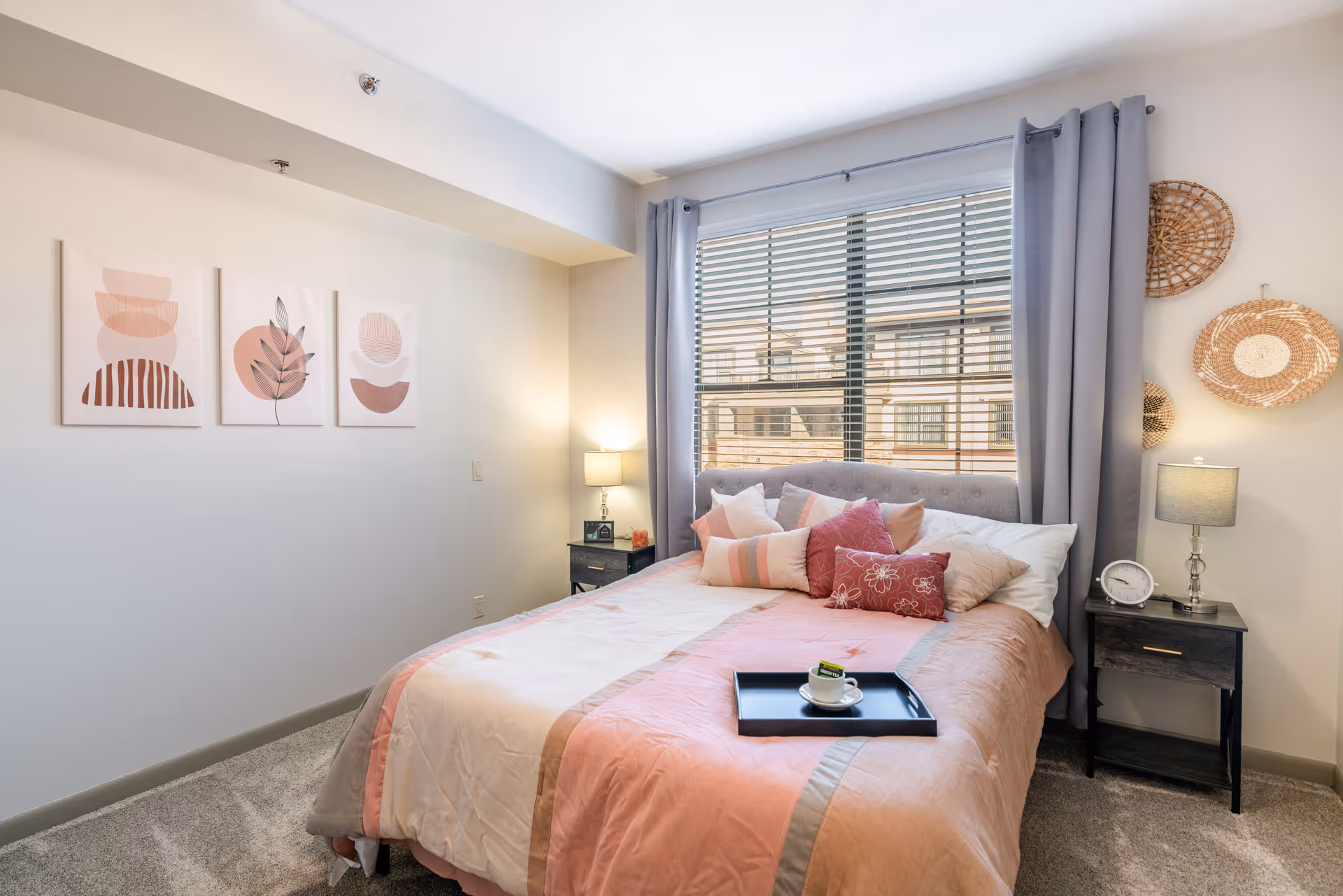A cozy bedroom with a neatly made bed featuring a pink and beige comforter and multiple decorative pillows. There is a black tray with a cup and saucer on the bed. The room has gray curtains framing a large window with blinds. On either side of the bed are black nightstands with lamps, a clock, and decorative items. The walls are adorned with three abstract art pieces and woven wall hangings. The floor is carpeted in a light gray color.