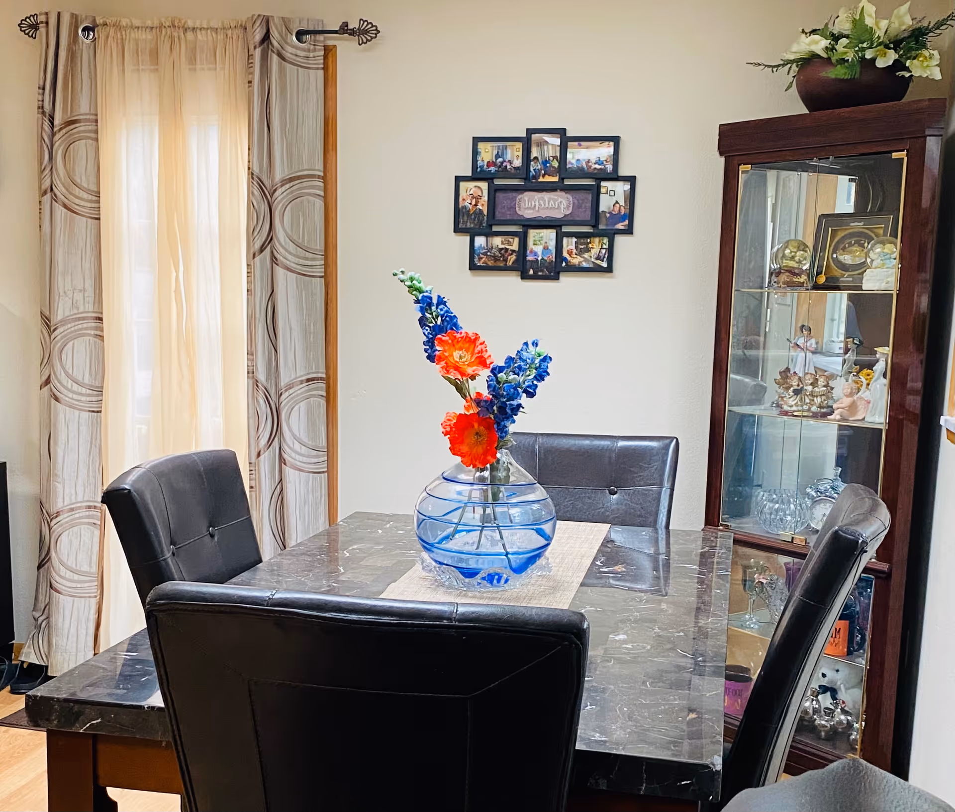 A dining room with a dark marble table surrounded by four black leather chairs. A clear glass vase with blue and red flowers is placed on a beige table runner in the center of the table. Behind the table, there is a window with beige curtains featuring circular patterns. On the wall above the table, there is a photo collage frame with multiple pictures. To the right, a wooden display cabinet with glass doors contains various decorative items and figurines.