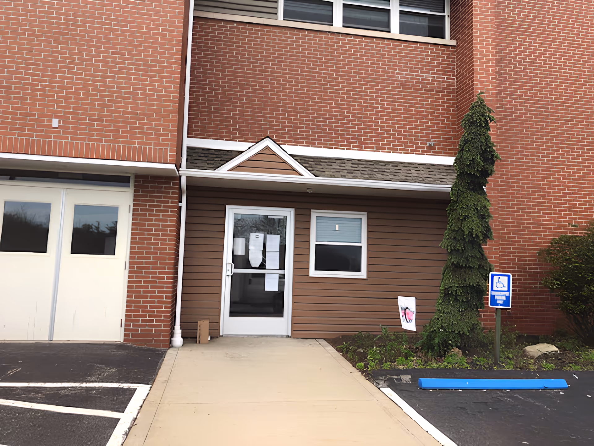 Entrance to a building with a glass door and a small window beside it, surrounded by brown siding and red brick walls. There is a sidewalk leading to the door, a small landscaped area with a tall, narrow tree, and a handicap parking sign with a blue parking block in front of it.