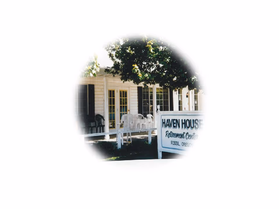 Exterior view of Haven House Retirement Center showing a white building with large windows and a porch area with white and black chairs. A tree provides shade over the porch and a sign in front reads 'Haven House Retirement Center Fossil, Oregon'.