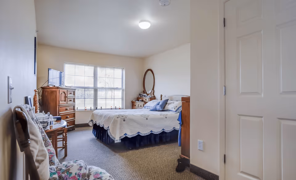A cozy bedroom in Devonshire Retirement Village featuring a bed with a white quilt and blue pillows, a wooden dresser with a TV on top, a wooden vanity with an oval mirror, and a window letting in natural light. There are two cushioned chairs with floral patterns near the entrance.