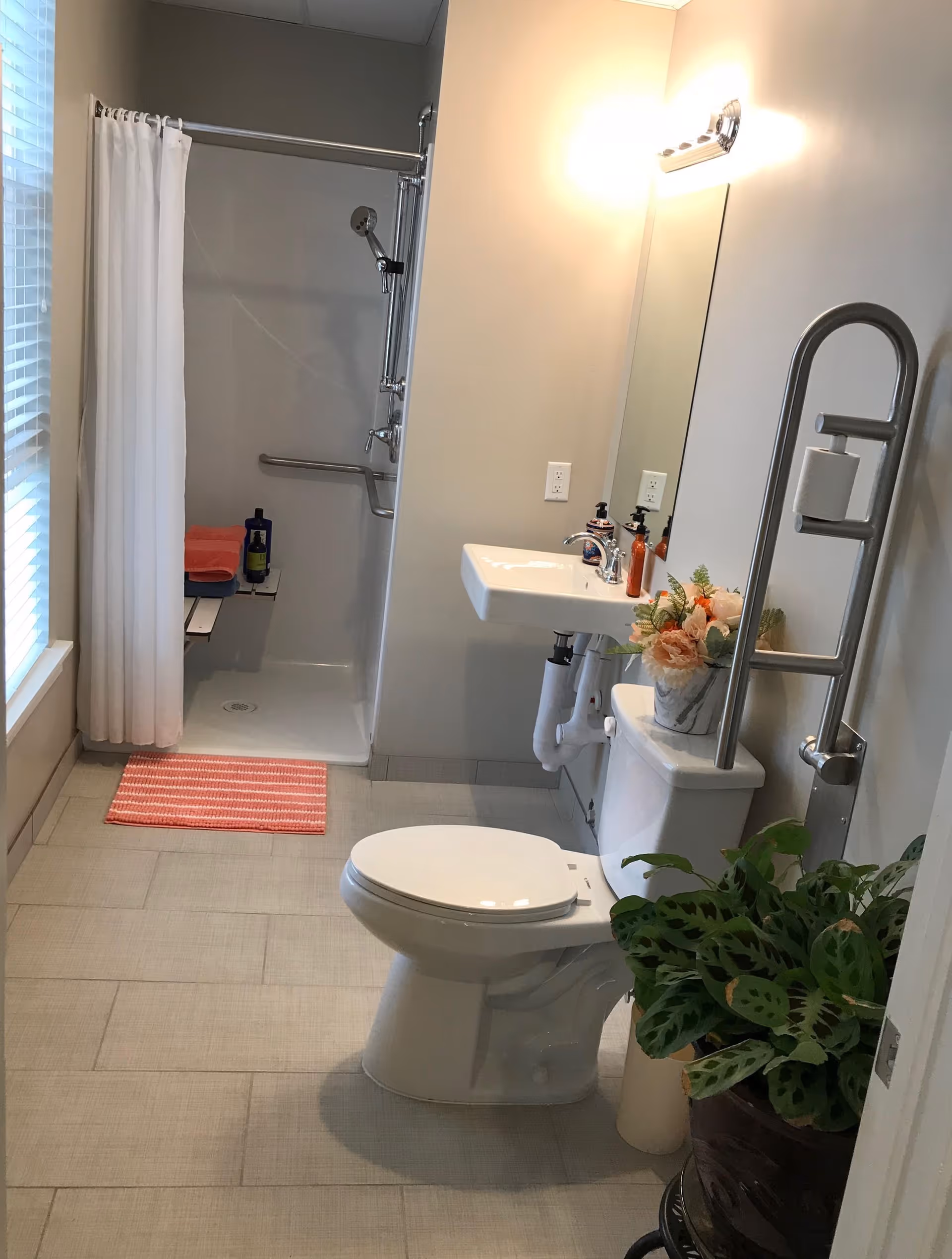 A clean, accessible bathroom with a white toilet, a wall-mounted sink with soap dispensers, a large mirror, and a walk-in shower with a white curtain and grab bars. There is a red bath mat on the floor and a potted plant near the toilet. The bathroom is well-lit with a window on the left side.