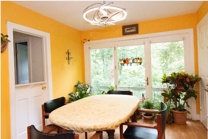 Sunlit dining room with an oval table covered in a cloth, chairs, potted plants, and large glass doors revealing a wooded view.