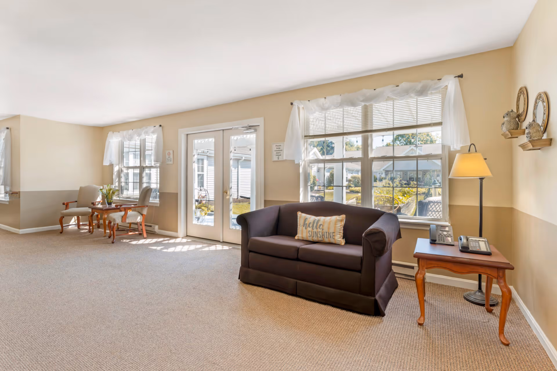 A bright and cozy common area in a senior living facility featuring a dark brown loveseat with a 'hello SUNSHINE' pillow, a wooden side table with two telephones and a floor lamp, large windows with white curtains letting in natural light, and a small seating area with two chairs and a wooden table near a glass door leading outside.