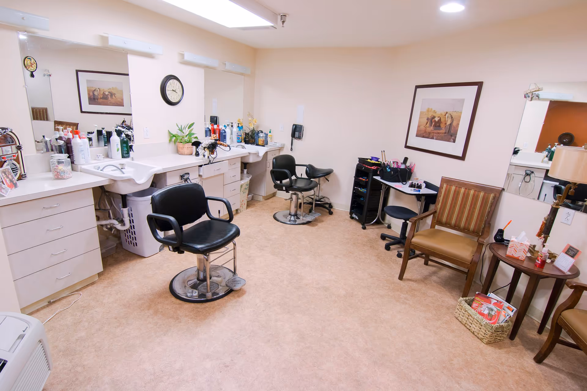 Interior of a senior living facility salon room with two black salon chairs in front of sinks and mirrors, various hair and beauty products on the counter, a clock on the wall, a framed picture, and additional seating with a small table holding tissues and magazines.