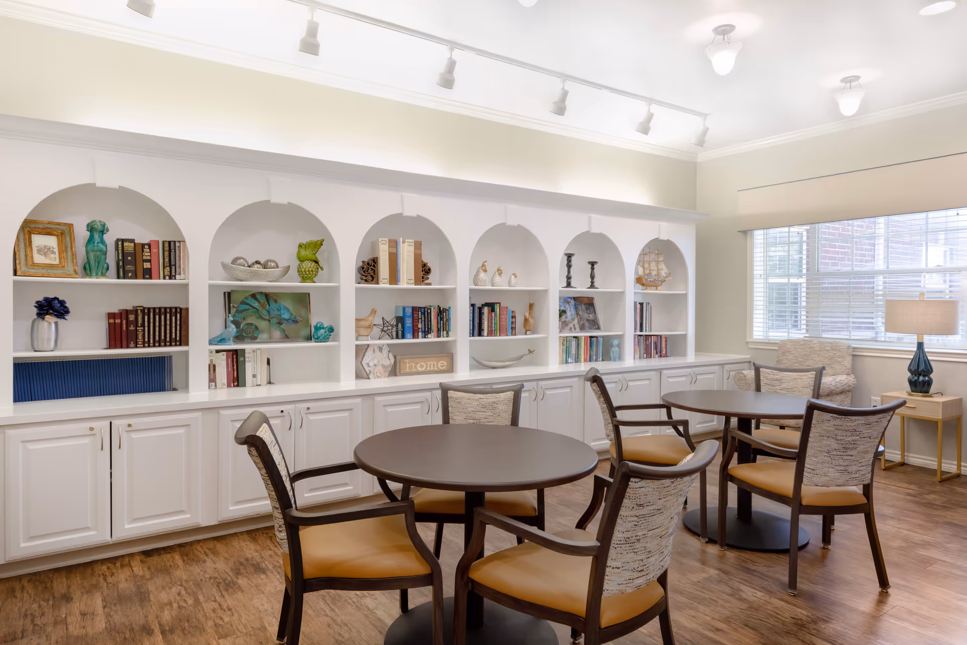 A well-lit room with two round tables surrounded by four cushioned chairs each. The back wall features white built-in shelves with decorative items, books, and art pieces. A large window with blinds and a side table with a lamp are visible on the right side. The floor has a wood finish.