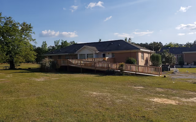 Single-story brick assisted living building with a wooden wheelchair ramp and a grassy lawn under a blue sky.
