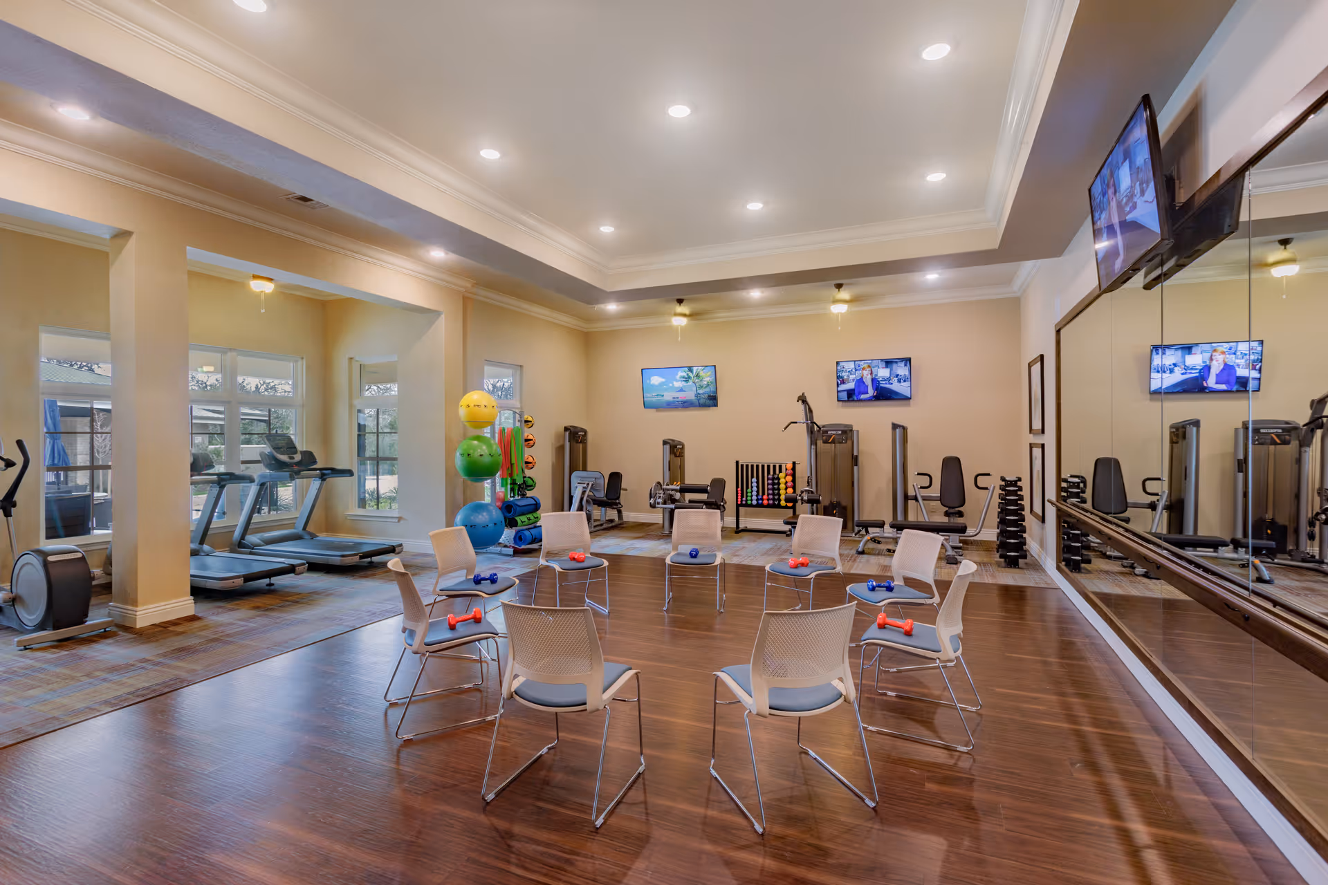 A fitness room with exercise equipment including treadmills, an elliptical machine, weight machines, and colorful exercise balls. There is a circle of chairs in the center of the room, each with small dumbbells placed on the seats. The room has large windows, a wall mirror, and two wall-mounted televisions.