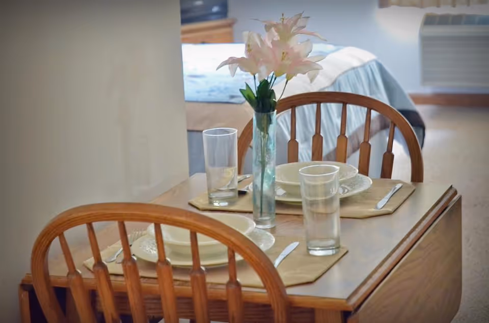 Small wooden dining table set for two with plates, glasses and a vase of flowers, with a bed visible in the background.