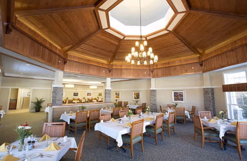 Bright dining room with an octagonal wood-paneled ceiling and chandelier, several tables set with white linens and place settings.
