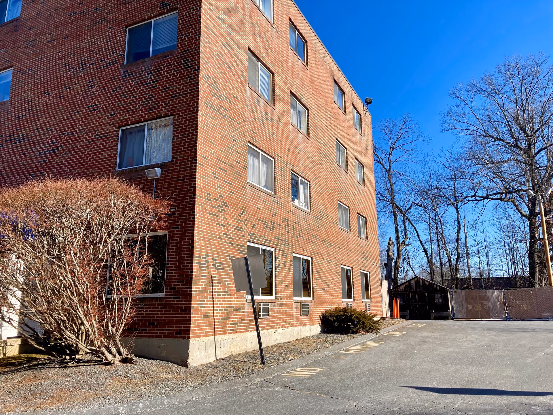 Exterior view of a multi-story brick building with several windows, a leafless bush near the corner, and a paved area with some trees in the background under a clear blue sky.