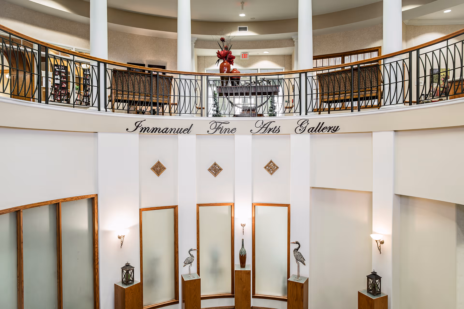 Interior view of Immanuel Fine Arts Gallery featuring a curved balcony with decorative railing, a table with a red vase and flowers on the upper level, and several art pieces displayed on pedestals below. The walls are white with wooden trim and decorative wall sconces providing lighting.