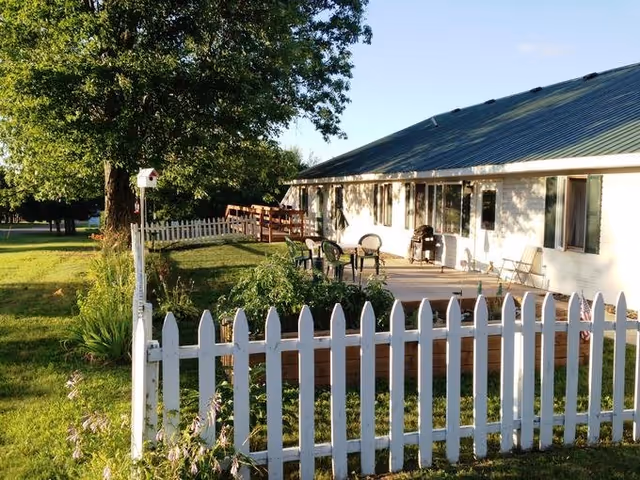 Outdoor view of a single-story building with a green metal roof and white exterior walls, surrounded by a white picket fence. There is a patio area with several chairs and a table, along with some greenery and trees in the background under a clear blue sky.