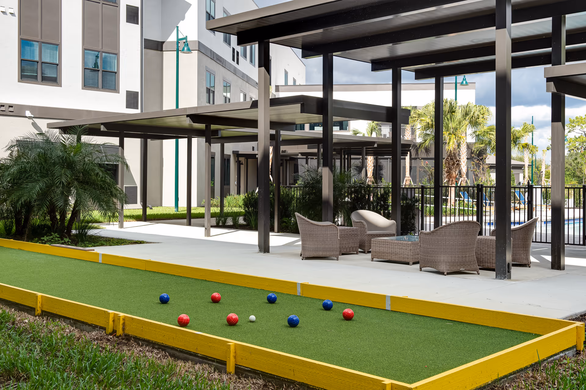 Outdoor bocce ball court with red and blue balls on a green surface bordered by yellow wooden edges, adjacent to a covered patio area with wicker chairs and a table, surrounded by palm trees and part of a multi-story building in the background.