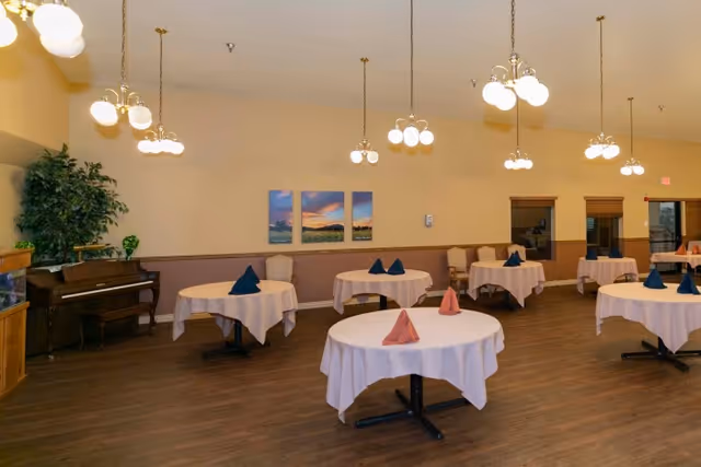 A spacious dining room with several round tables covered with white tablecloths and neatly folded napkins in blue and pink. The room features wooden flooring, multiple hanging light fixtures with globe-shaped bulbs, a piano against one wall, and a large green plant beside it. Three landscape paintings hang on the beige wall, and there are windows with wooden blinds on the right side.