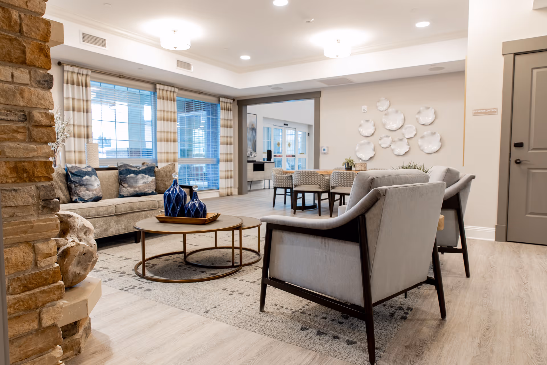 A cozy senior living facility common area with a stone fireplace on the left, a beige sofa with blue patterned pillows, two gray armchairs, a round wooden coffee table with decorative blue vases, and a dining table with chairs in the background. The room has large windows with striped curtains and light wood flooring.