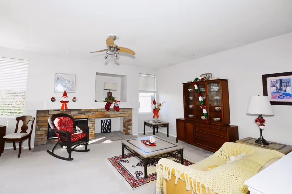 A bright living room decorated for Christmas with a stone fireplace adorned with stockings and festive decorations. The room features a wicker rocking chair with a red cushion, a wooden cabinet with dishes and a garland, a coffee table on a patterned rug, a yellow couch with a knitted throw, and a table lamp. Two windows with blinds allow natural light to fill the space.