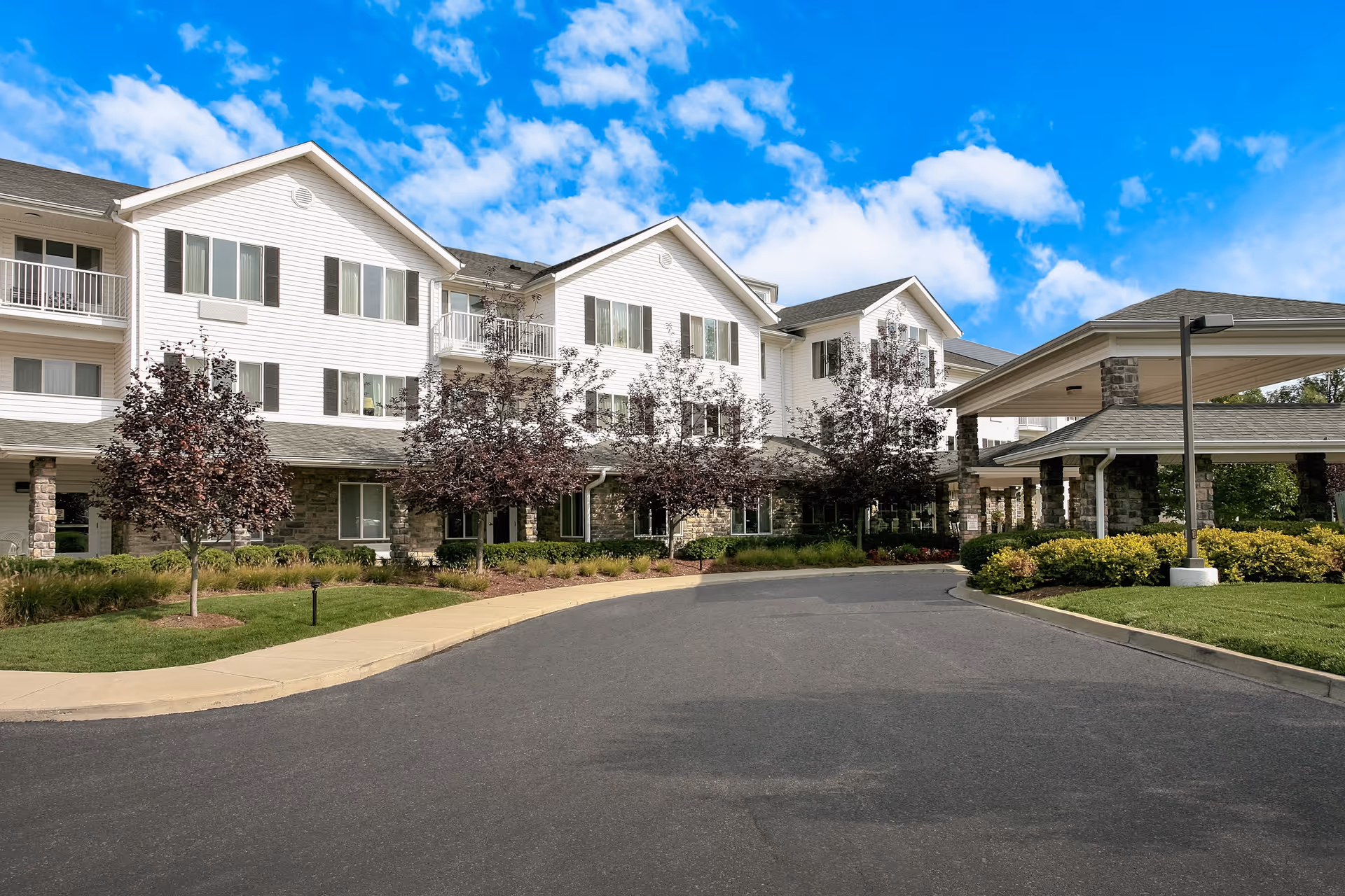 Exterior view of a multi-story senior living facility building with white siding and stone accents. The building has multiple windows and balconies, with a covered entrance driveway surrounded by landscaped bushes and small trees under a partly cloudy blue sky.