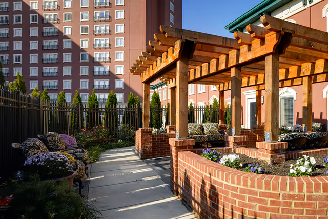Sunlit outdoor courtyard featuring a wooden pergola, brick planters, cushioned seating, and a multi-story building in the background.