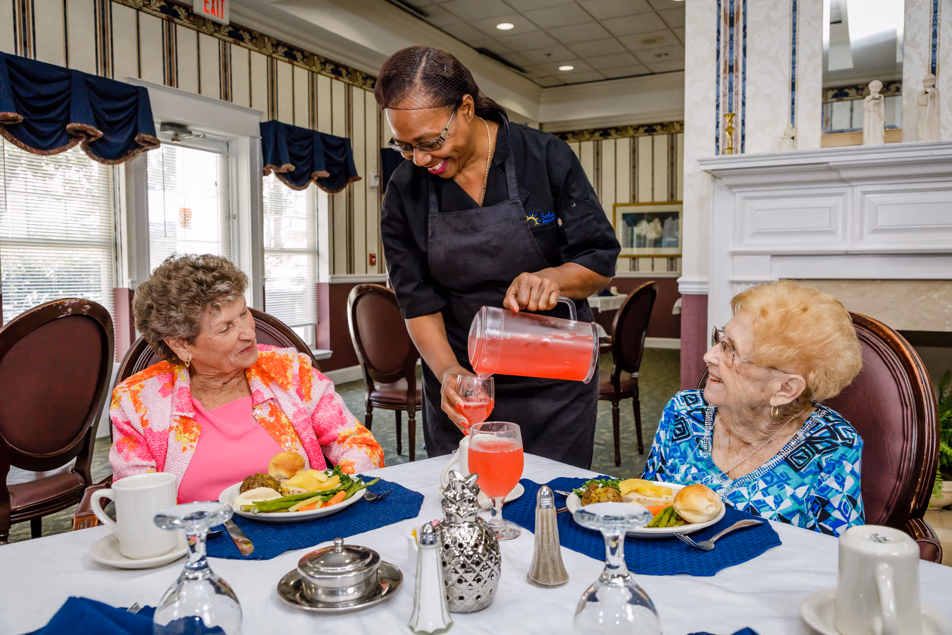 A server pours a pink beverage into a glass for two elderly women seated at a dining table in a dining room.