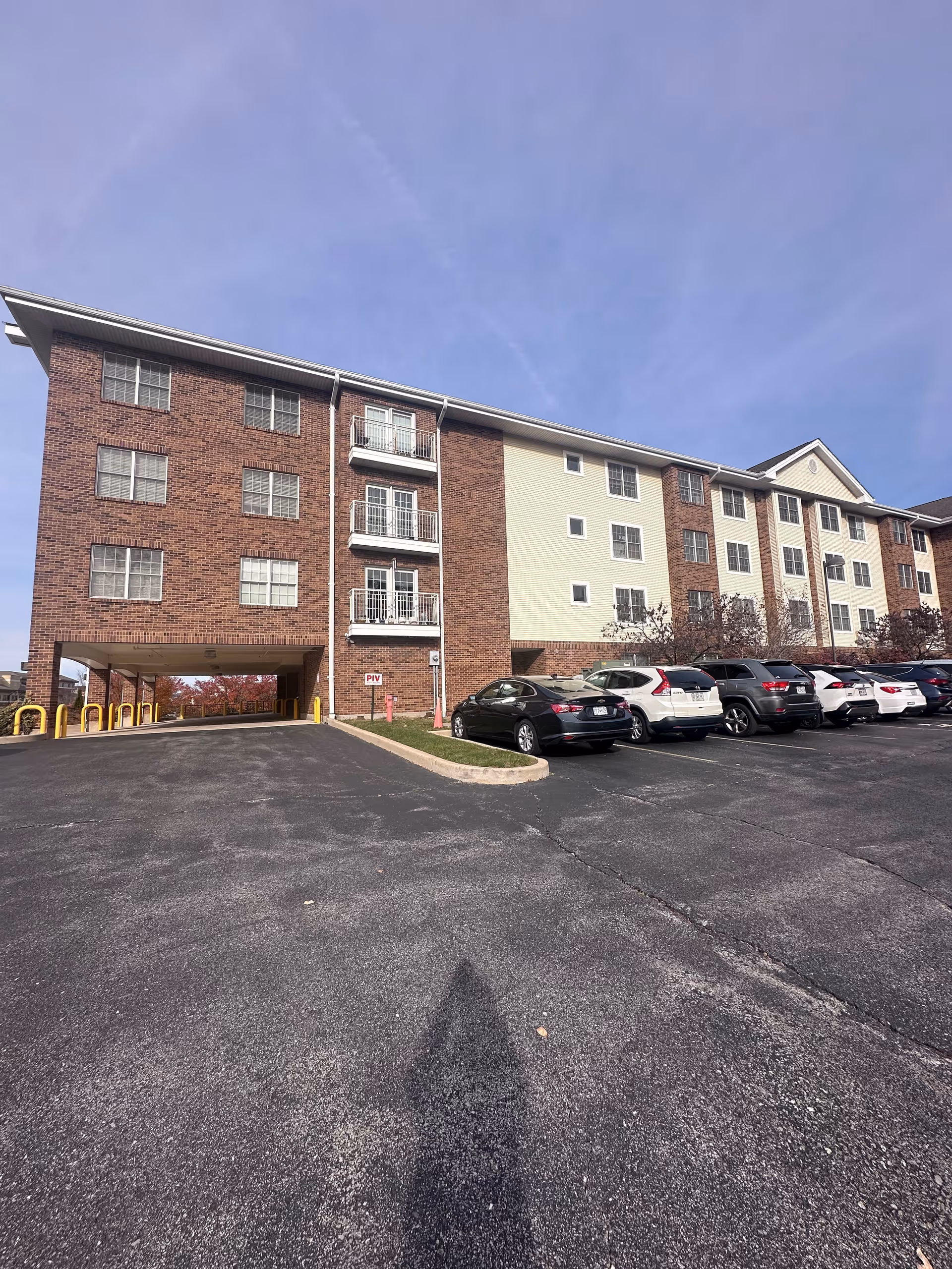 Exterior view of a four-story brick-and-siding apartment building with balconies and parked cars in a large asphalt parking lot.